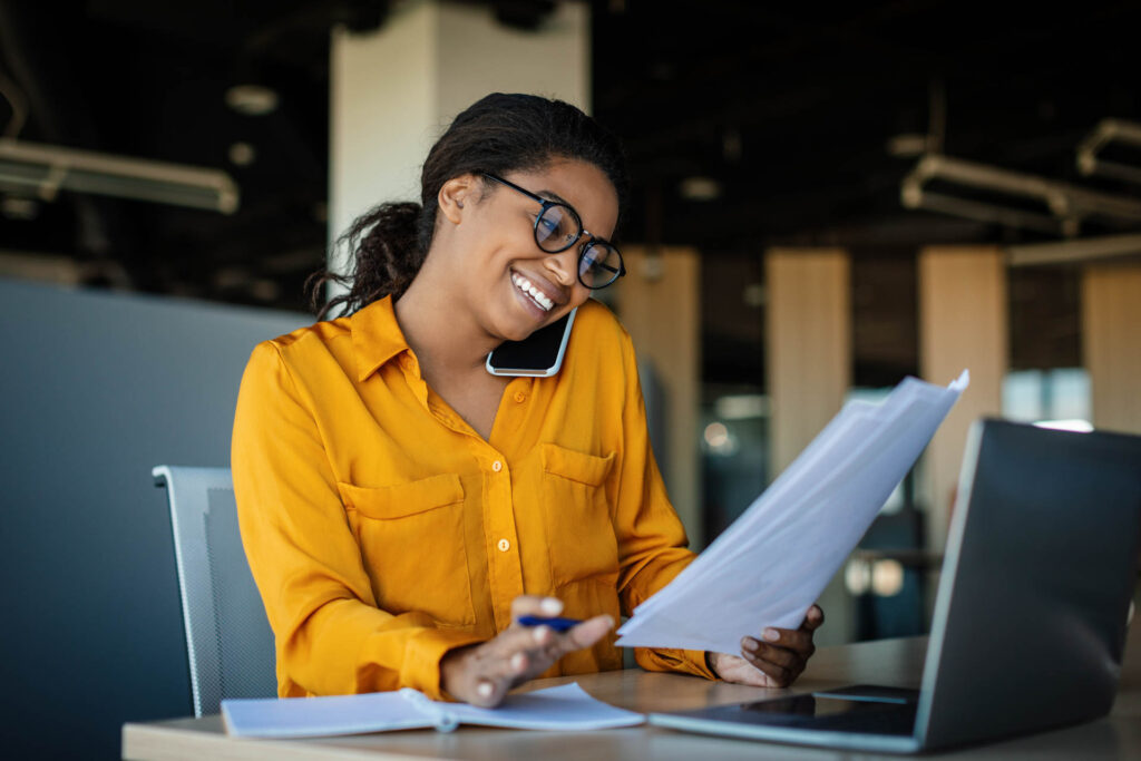 Corporate communication. Happy black female manager working with papers and talking on mobile phone, reading financial documents while sitting at workplace in office